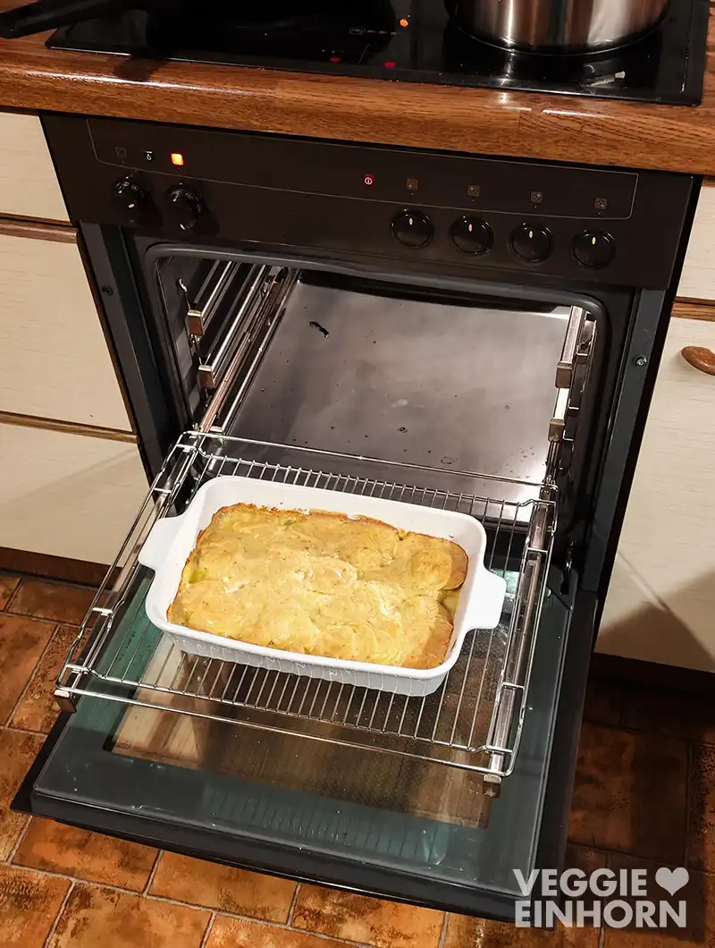 Geöffneter Backofen, darin eine weiße Auflaufform mit goldbraunem veganem Kartoffelgratin auf dem Rost.