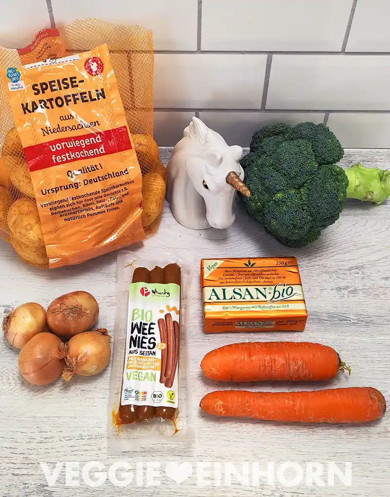 Zutaten für einen veganen Eintopf liegen auf einer hellen Arbeitsfläche, darunter Kartoffeln im Netz, frischer Brokkoli, Zwiebeln, vegane Wiener Würstchen aus Seitan, Alsan-Bio-Margarine, zwei Möhren und eine Einhornfigur im Hintergrund.