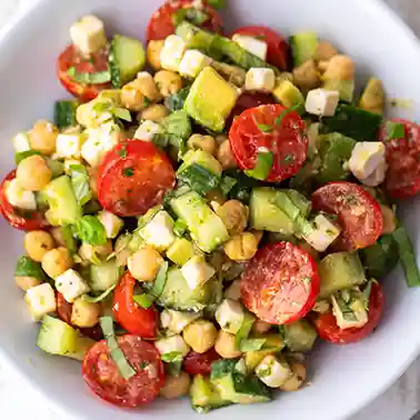 Veganer Salat mit Kichererbsen, veganem Feta, Avocado, Kirschtomaten, Salatgurke und Kräutern auf einem weißen Teller.