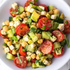 Draufsicht auf einen veganen Kichererbsensalat mit Avocado, Gurke, Cherrytomaten, veganem Feta, Petersilie und Basilikum auf einem weißen Teller.