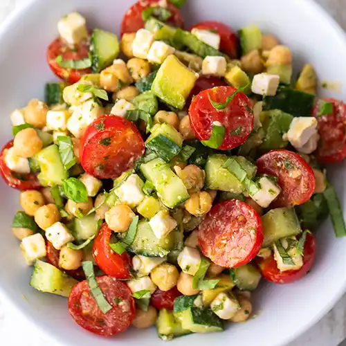 Draufsicht auf einen veganen Kichererbsensalat mit Avocado, Gurke, Cherrytomaten, veganem Feta, Petersilie und Basilikum auf einem weißen Teller.
