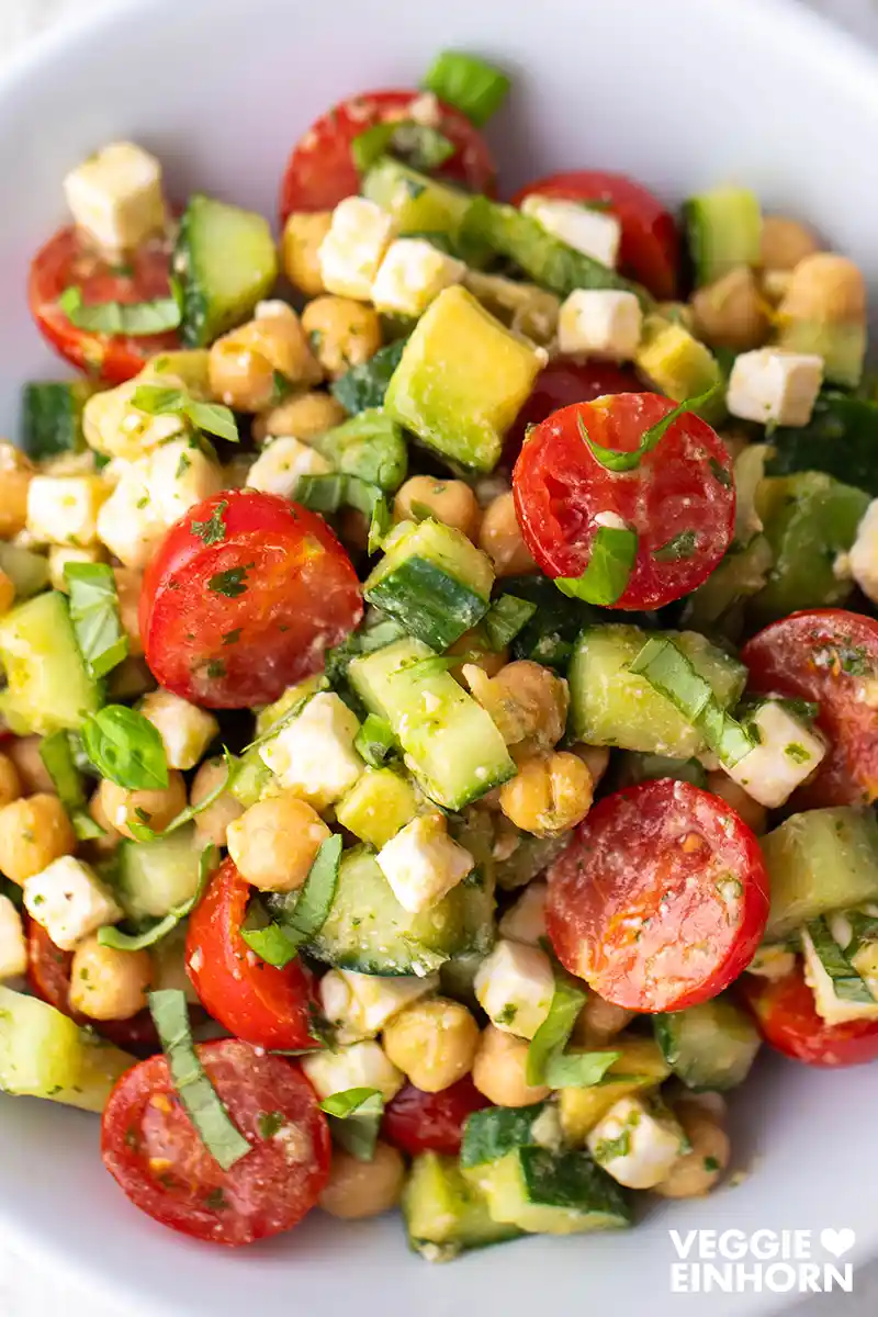 Draufsicht auf einen bunten Kichererbsensalat mit Avocado, Gurke, Cherrytomaten, veganem Feta und frischen Kräutern auf einem weißen Teller.