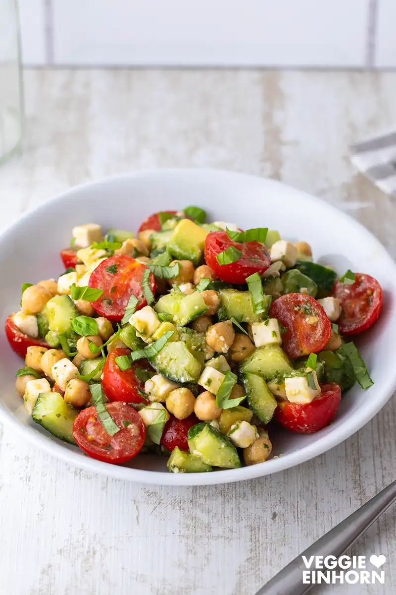 Frischer Kichererbsensalat mit Avocado, Gurke, Cherrytomaten, veganem Feta und Basilikum auf einem weißen Teller auf hellem Untergrund.