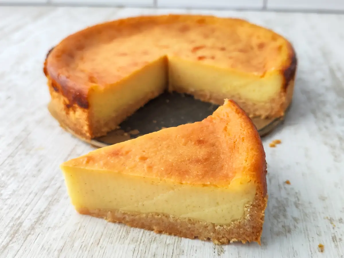 Ein Stück kleiner veganer Käsekuchen liegt vor dem angeschnittenen Kuchen auf einer weißen Holzoberfläche.