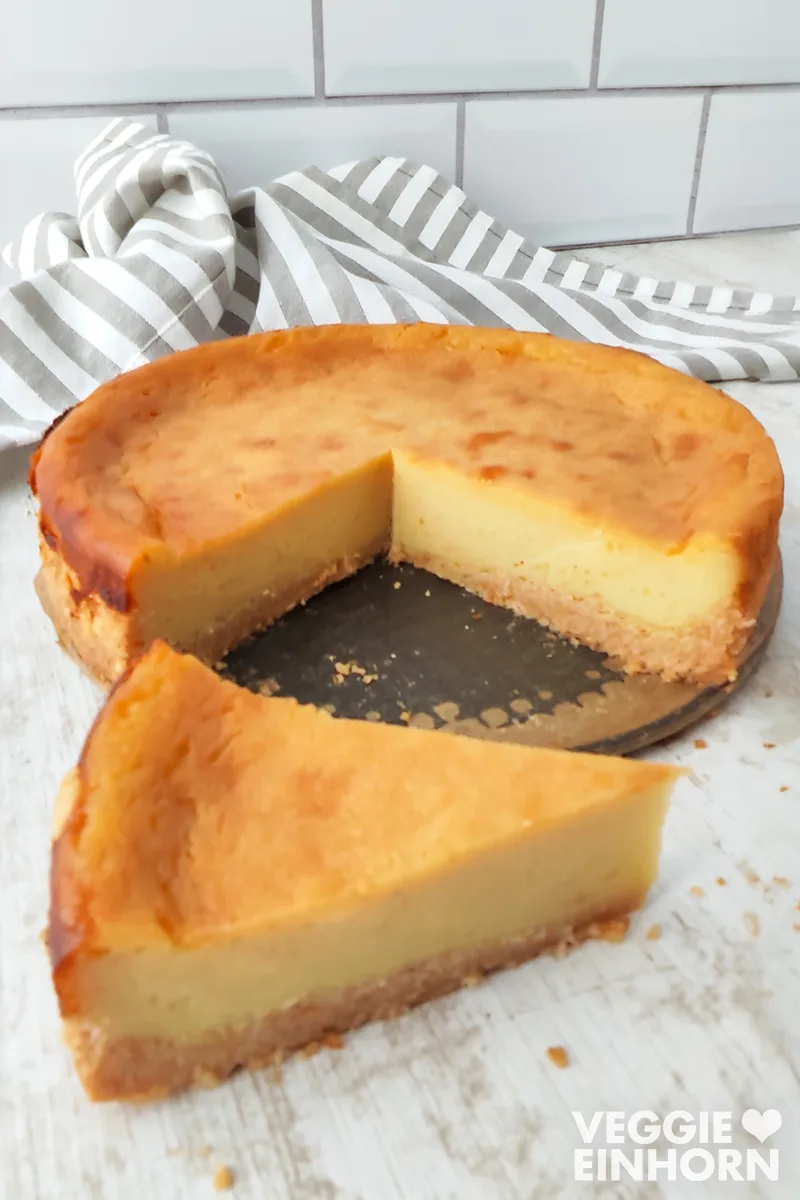 Kleiner veganer Käsekuchen auf heller Holzoberfläche, im Hintergrund ein gestreiftes Küchentuch.