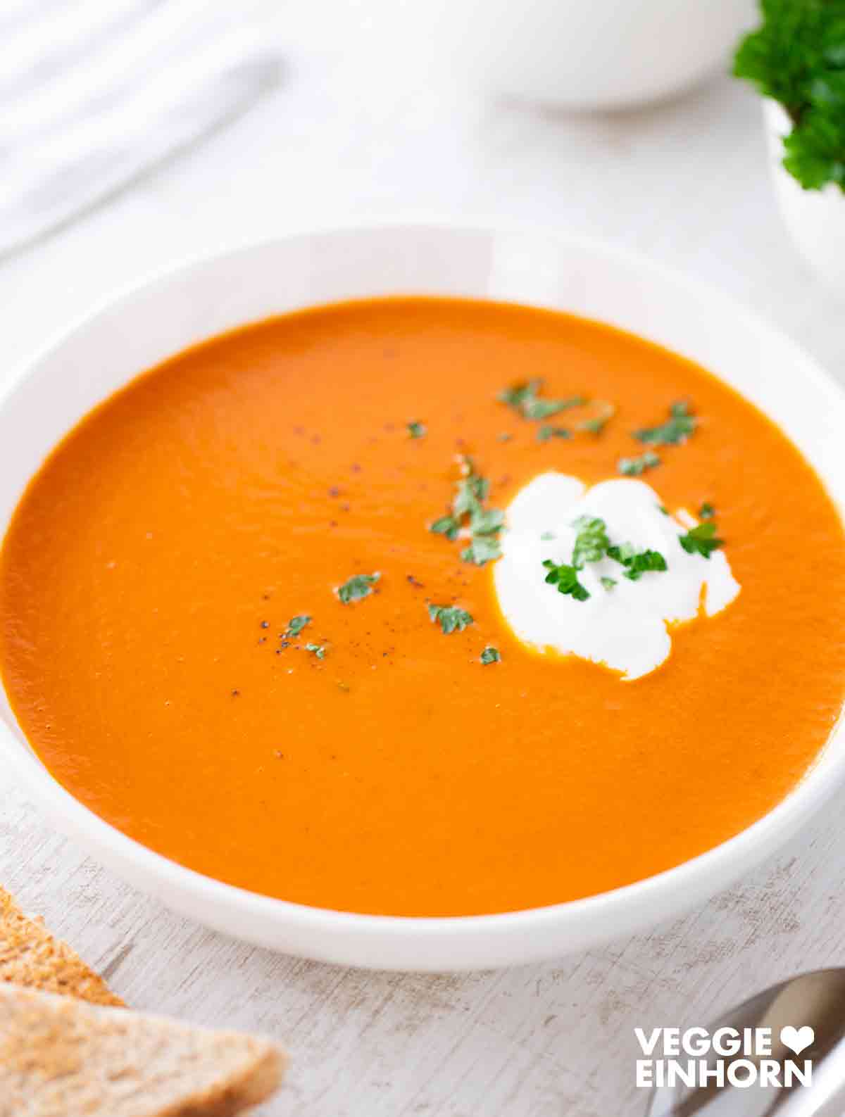 Weiße Schüssel mit veganer Tomatensuppe, garniert mit einem Klecks Kokosmilch und gehackter Petersilie.