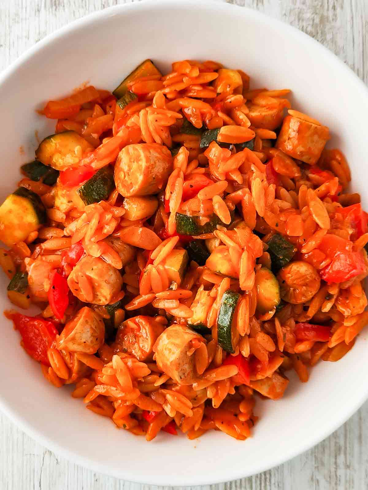 Draufsicht auf einen weißen Teller mit einer Gemüsepfanne aus Kritharaki-Nudeln, veganen Bratwurststücken, Zucchini und Paprika in einer Tomatensoße.