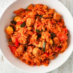 Weißer Teller mit einer Portion von einer Kritharaki-Pfanne mit Tomatensoße, darin Stücke von veganen Bratwürstchen, Paprika und Zucchini,