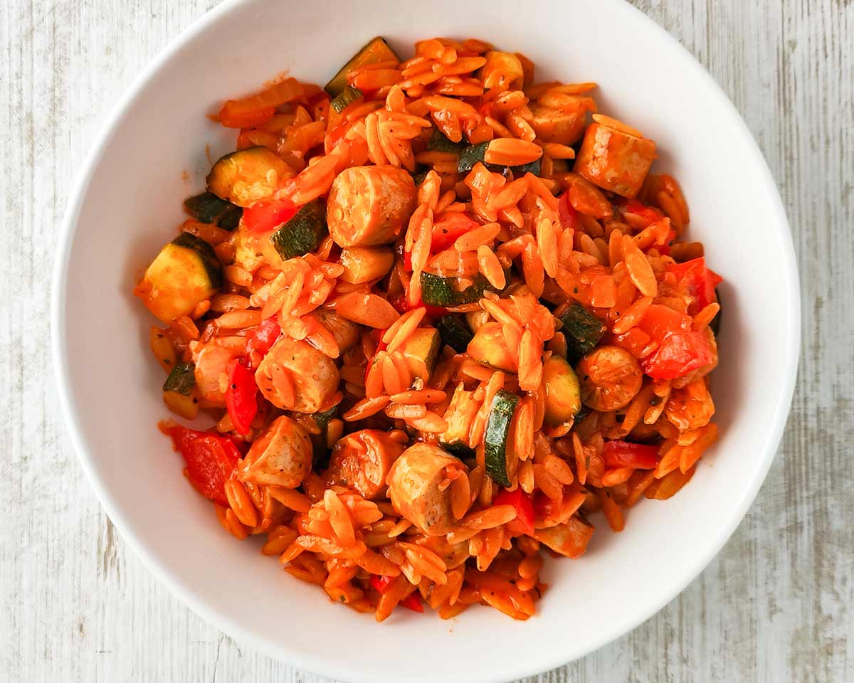 Weißer Teller mit einer Portion von einer Kritharaki-Pfanne mit Tomatensoße, darin Stücke von veganen Bratwürstchen, Paprika und Zucchini,