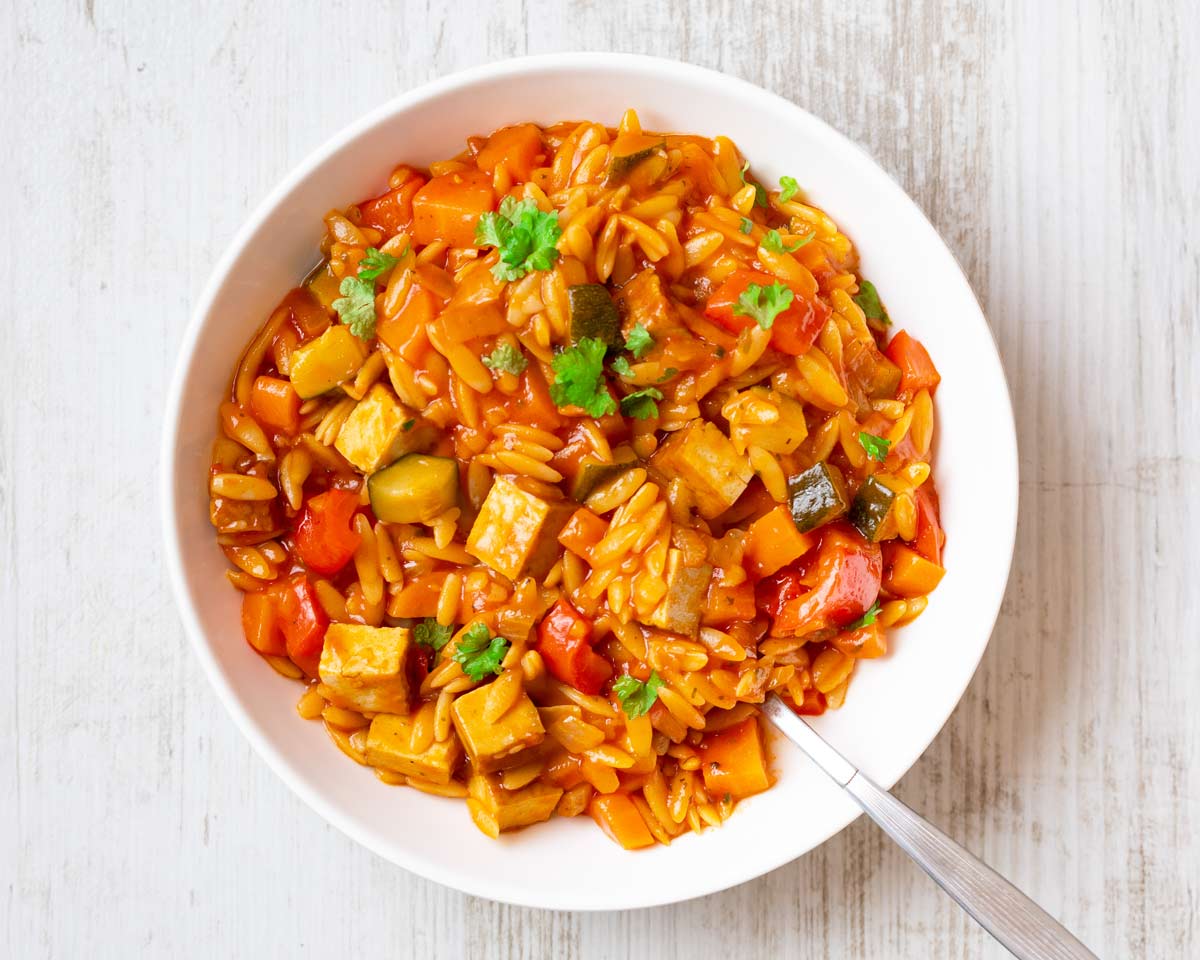 Vegane Kritharaki-Pfanne mit Räuchertofu und Gemüse in einem weißen Teller, garniert mit frischer Petersilie.