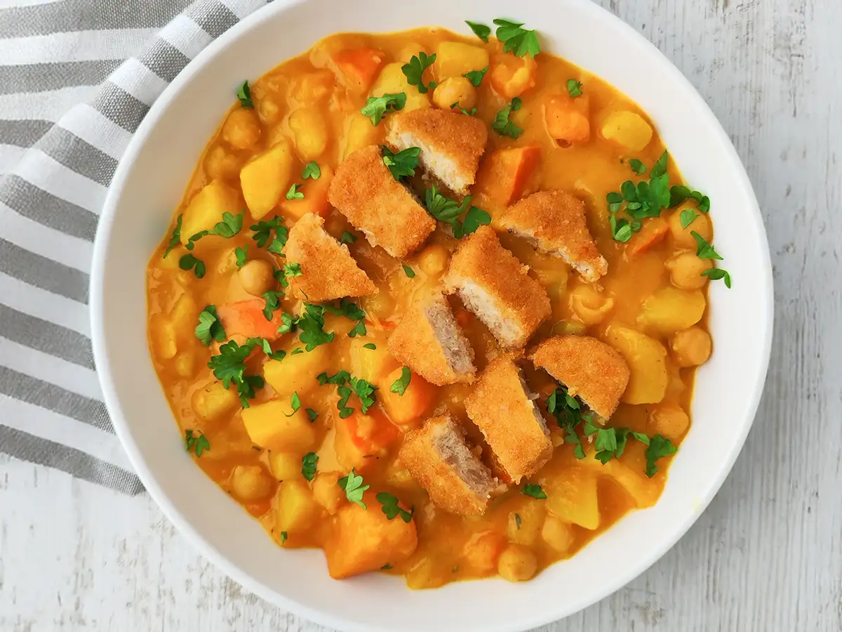 Ein Teller veganer Kürbis-Kichererbsen-Eintopf mit Kartoffeln und veganen Nuggets, garniert mit frischer Petersilie.