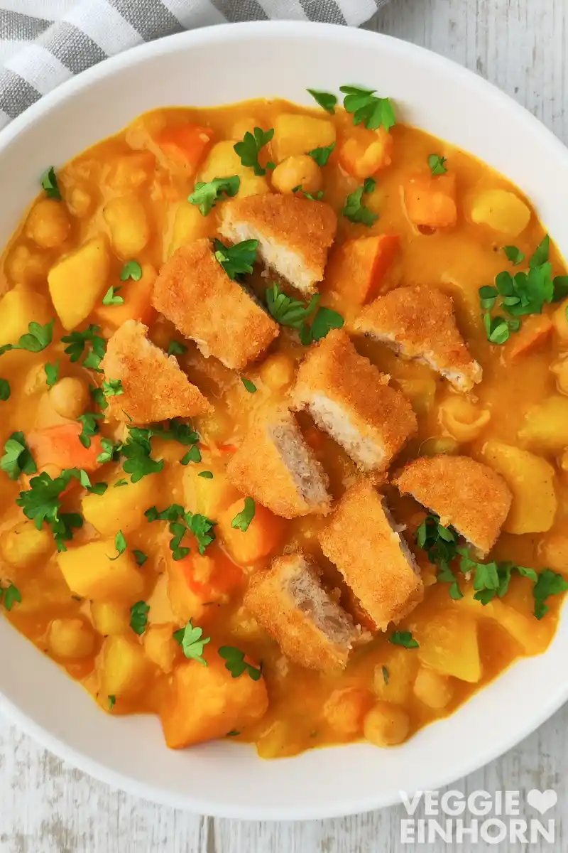 Teller mit Kürbis-Kichererbsen-Eintopf mit Kartoffeln mit goldbraunen veganen Nuggets, garniert mit Petersilie.
