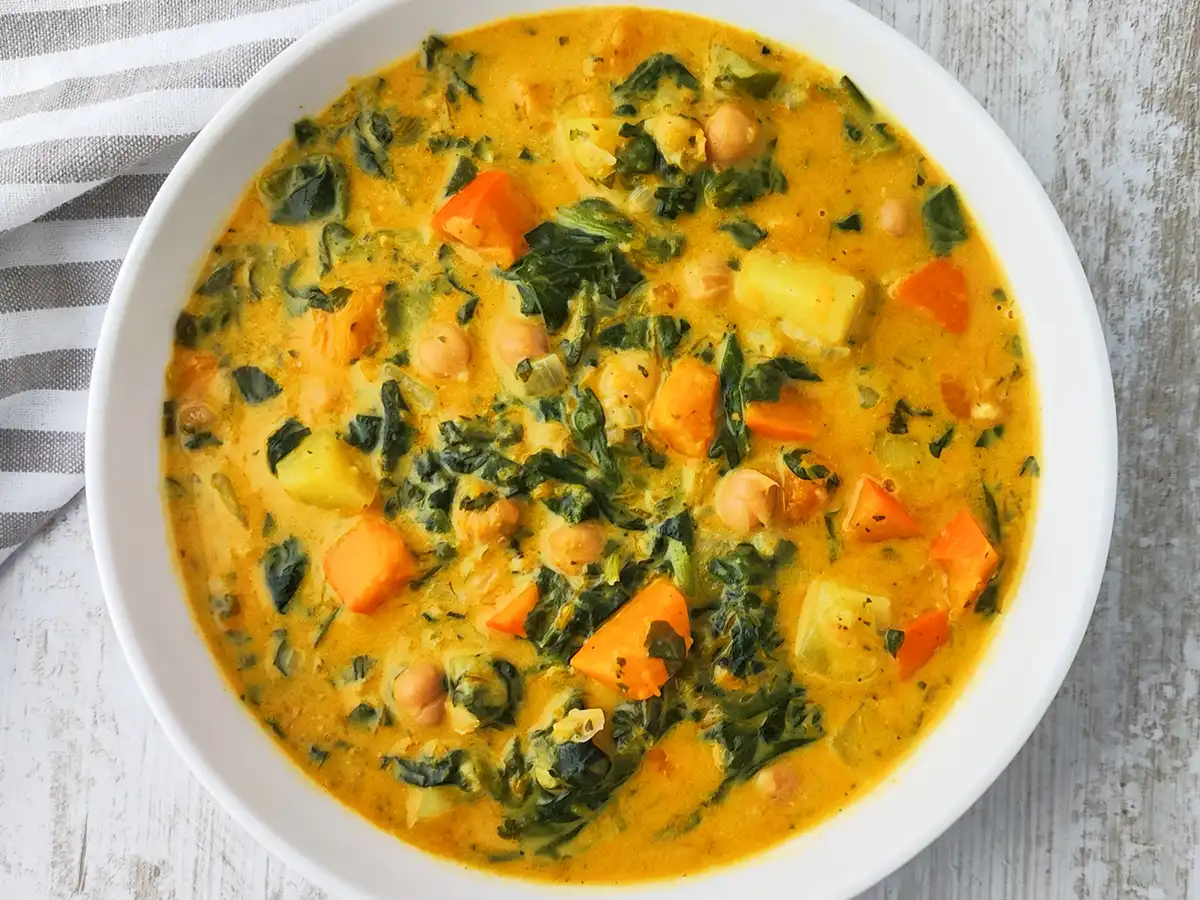 Eine weiße Schüssel ist mit einem cremigen Kürbis Kichererbsen Eintopf gefüllt, in dem Spinat, Kartoffeln und orangefarbene Kürbisstücke zu sehen sind.
