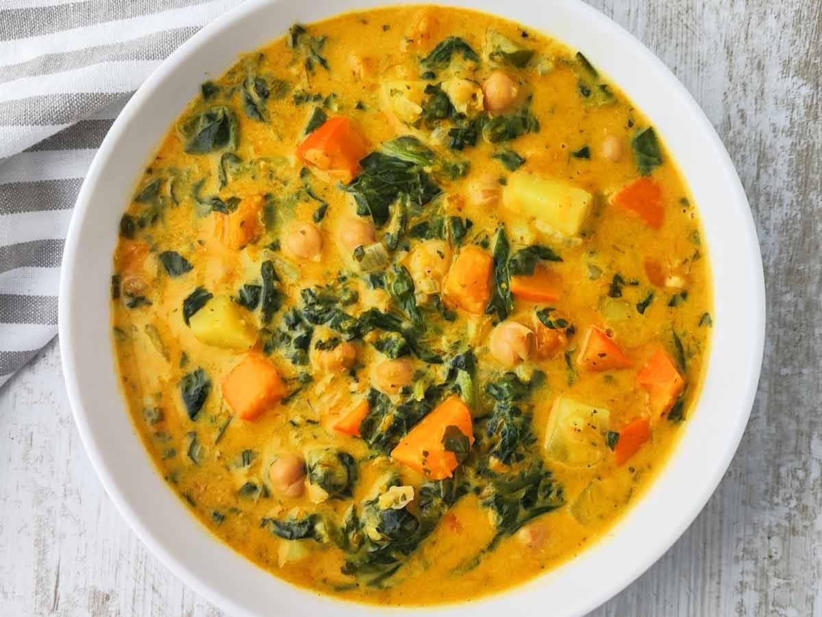 Eine Nahaufnahme zeigt einen cremigen Kürbis Kichererbsen Eintopf mit Spinat, Kartoffeln und Kürbisstücken in einer weißen Schüssel auf einer hellen Oberfläche.