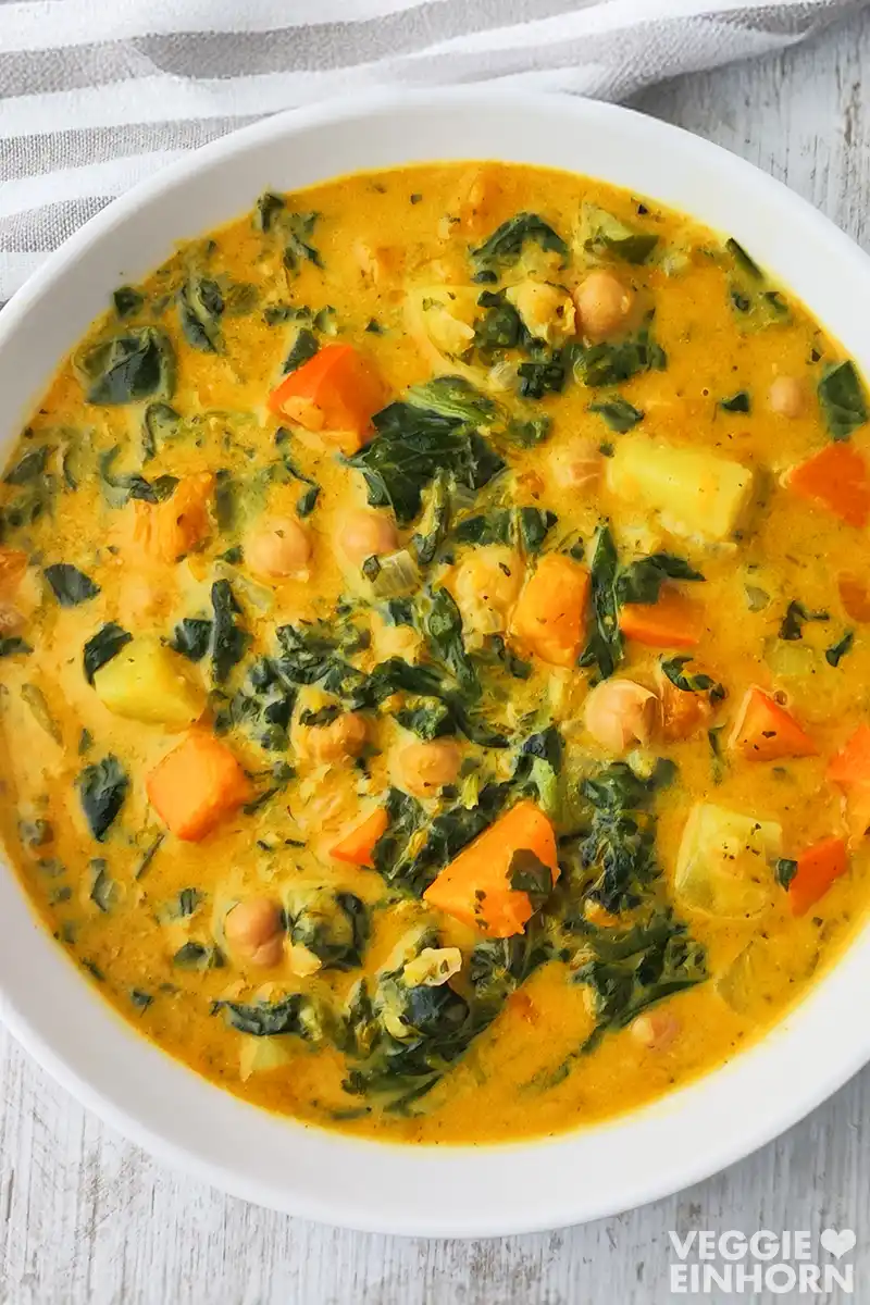 Ein weißer Teller ist mit einem cremigen veganen Kürbis Kichererbsen Eintopf gefüllt, in dem Spinat und Kartoffeln zu sehen sind. Daneben liegt ein gestreiftes Küchentuch.