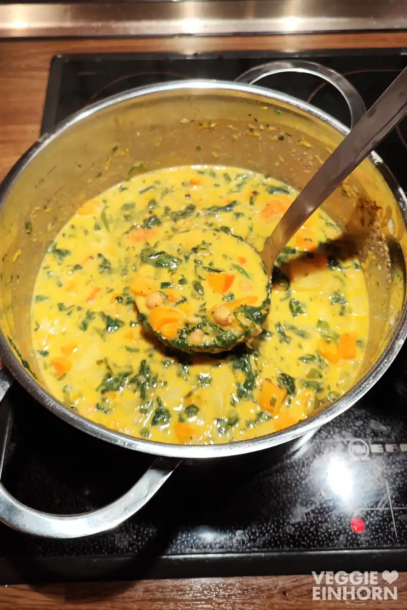 Ein großer Topf steht auf dem Herd und ist gefüllt mit einem cremigen Kürbis Kichererbsen Eintopf mit Spinat, in dem ein Schöpflöffel steckt.