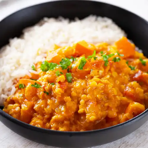 Cremiges Linsencurry mit Kürbisstücken, roten Linsen und Reis in einem schwarzen Teller, garniert mit Petersilie.