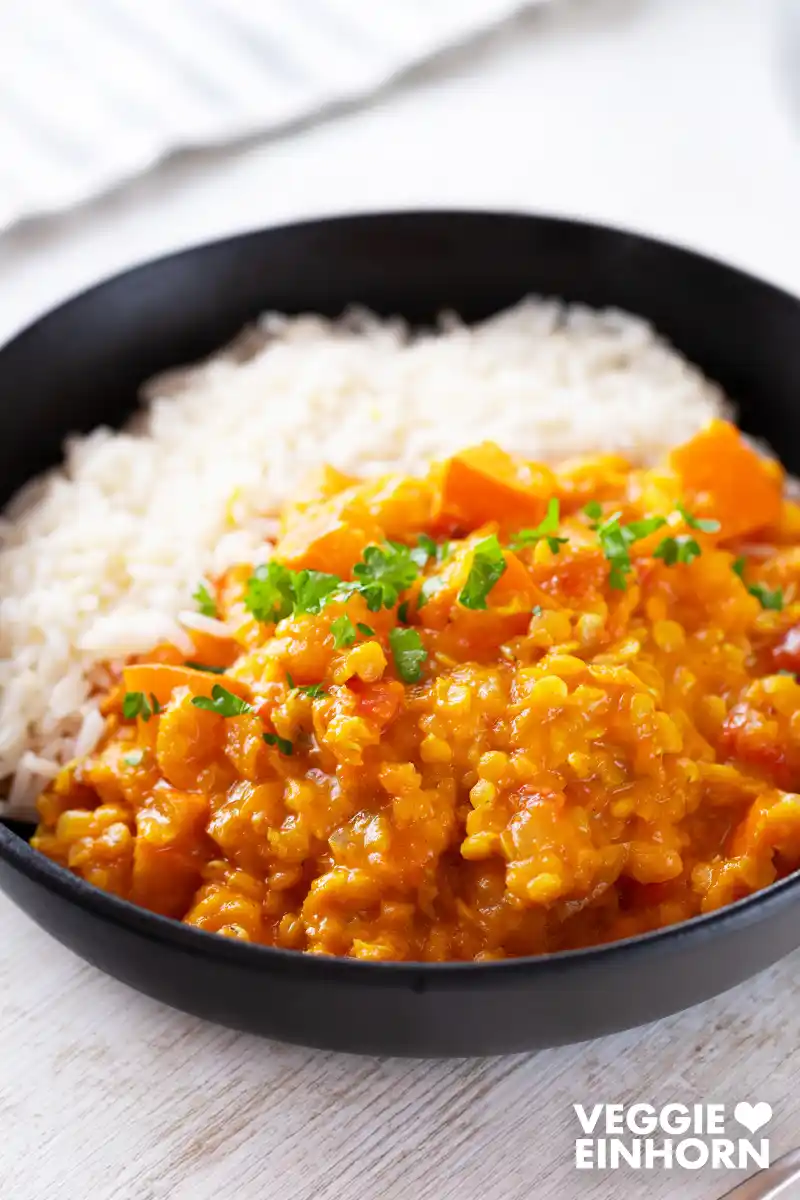 Veganes Kürbis-Linsen-Curry mit Reis, serviert in einem schwarzen Teller und bestreut mit frischer gehackter Petersilie.