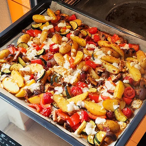 Buntes Ofengemüse mit Kartoffeln, Zucchini, Tomaten und Paprika auf einem Backblech, überbacken mit veganem Feta.