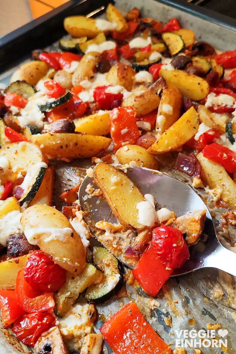 Nahaufnahme von mediterranem Ofengemüse auf einem Blech mit Kartoffelspalten, Paprika, Zucchini, Tomaten, Champignons und veganem Feta, teilweise bereits mit einem Servierlöffel herausgehoben.