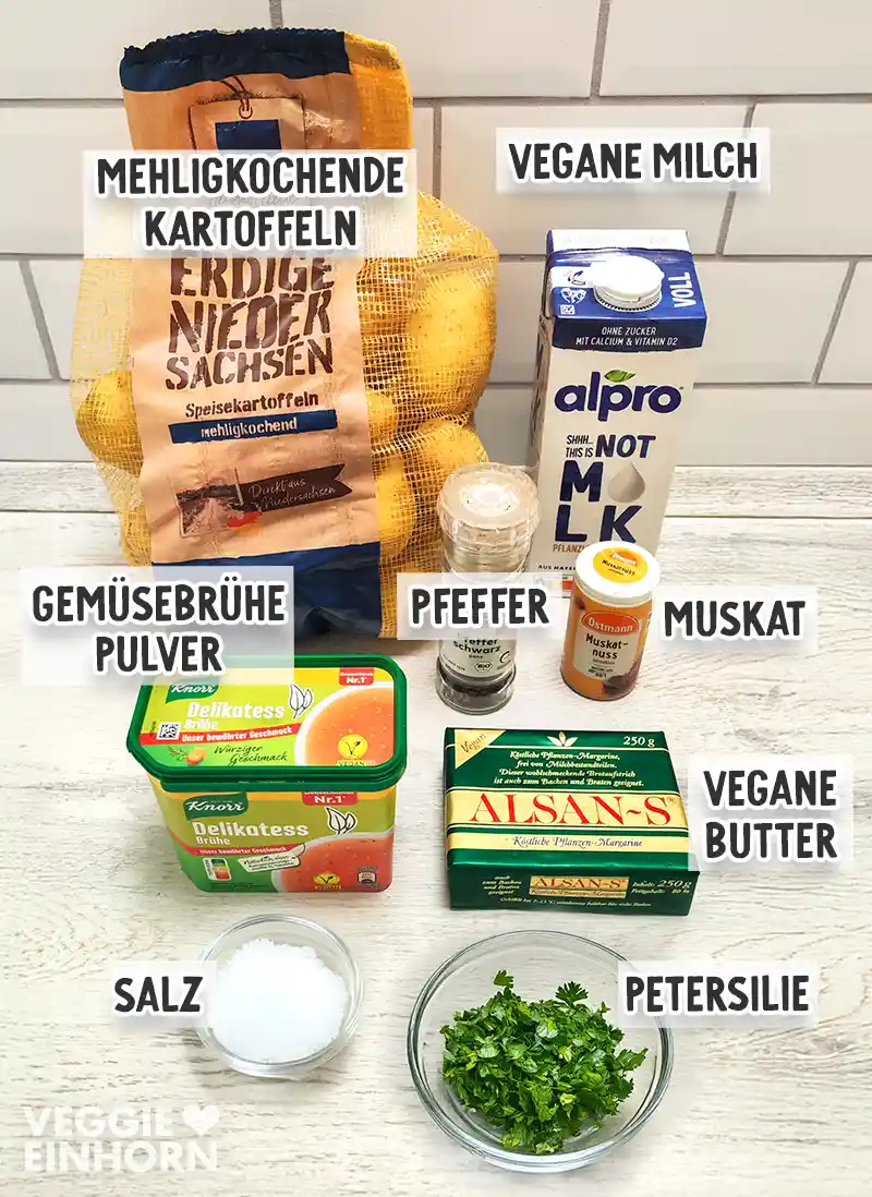 Zutaten für veganes Kartoffelpüree mit mehligkochenden Kartoffeln, veganer Milch, Gemüsebrühepulver, Pfeffer, Muskat, veganer Butter, Salz und gehackter Petersilie auf einem hellen Tisch.