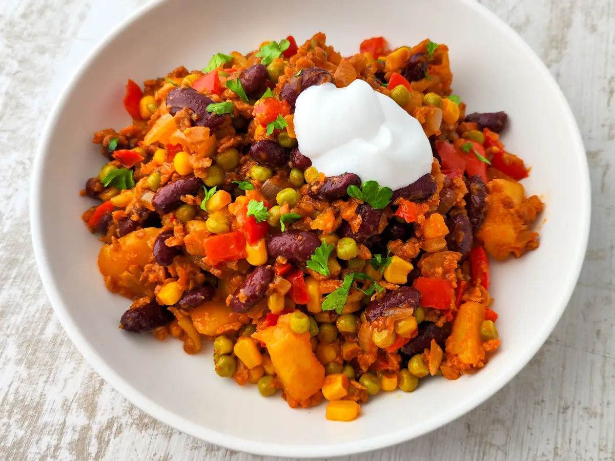 Vegane mexikanische Kartoffelpfanne mit Kidneybohnen, Mais, Erbsen, Paprika und veganem Hack, serviert auf einem weißen Teller und mit einem Klecks veganer Crème fraîche garniert.