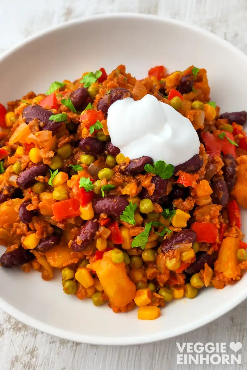 Vegane mexikanische Kartoffelpfanne mit Kidneybohnen, Mais, Erbsen, Paprika und veganem Hack, serviert auf einem weißen Teller, mit frischer Petersilie und einem Klecks veganer Crème fraîche garniert.