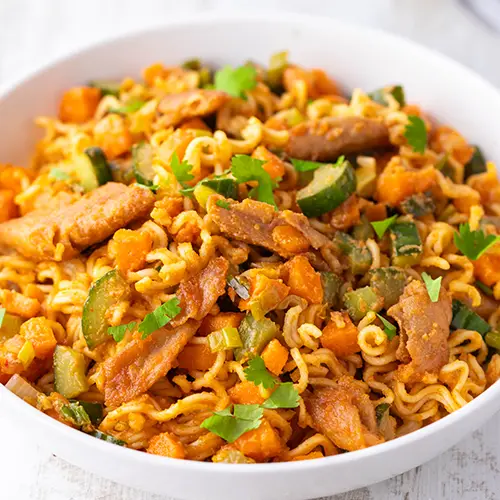 Weißer Teller mit Mie-Nudeln, veganem Hähnchen, Gurke und Möhren. Die bunte Nudelpfanne ist mit frischer Petersilie bestreut und steht auf hellem Untergrund.