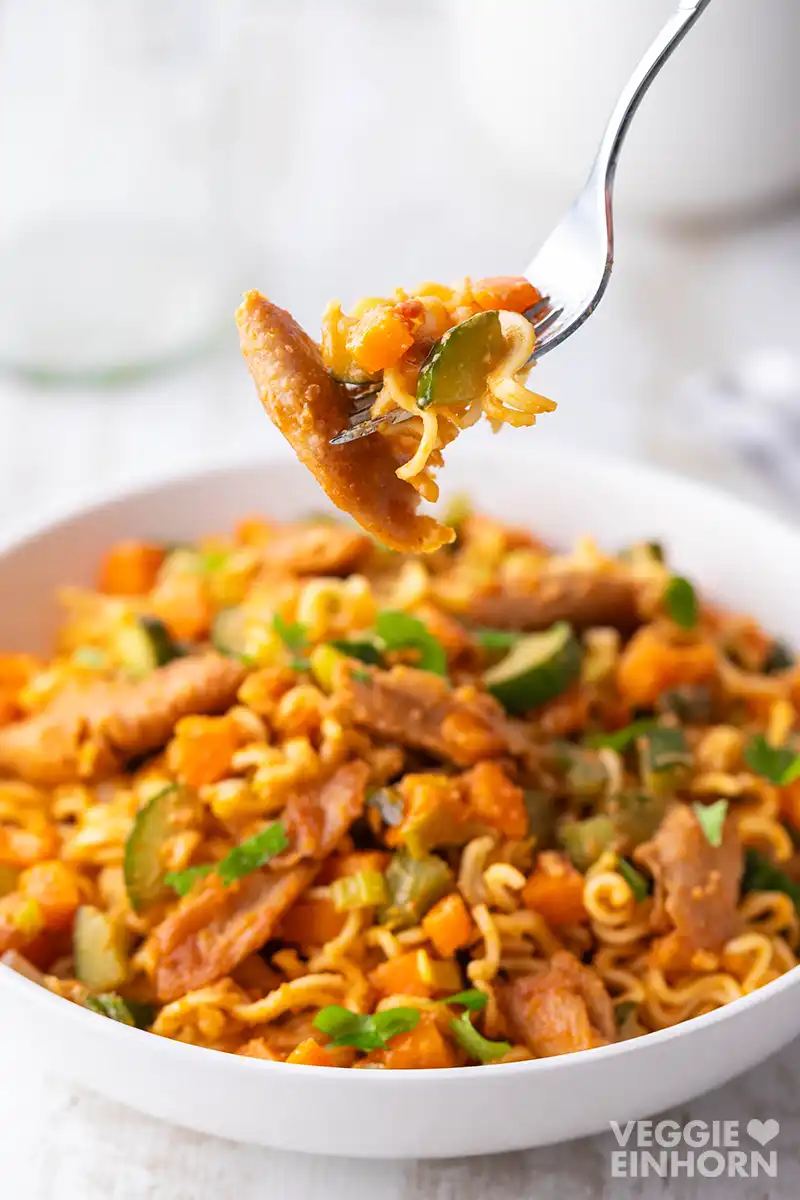 Eine Gabel mit Mie-Nudeln, Gurke, Möhre und veganem Hähnchenstück wird über einem Teller mit dem Rest der Mahlzeit gehalten.
