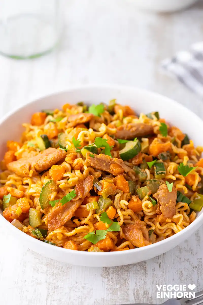 Weißer Teller mit Mie-Nudeln, veganem Hähnchen, Gurke, Möhren und Frühlingszwiebeln auf hellem Untergrund. Das Gericht ist mit frischer Petersilie bestreut.