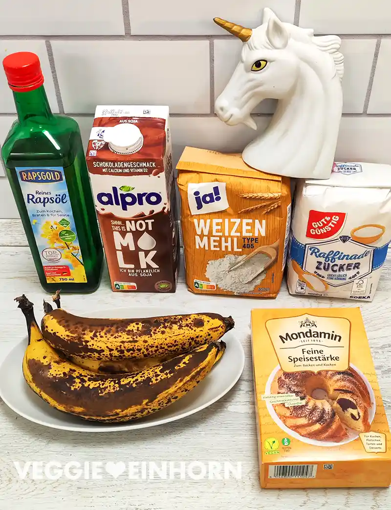 Mehrere Backzutaten stehen auf einer Küchenarbeitsplatte, darunter Rapsöl, Schoko-Sojamilch, Mehl, Zucker, Speisestärke und sehr reife Bananen auf einem Teller.