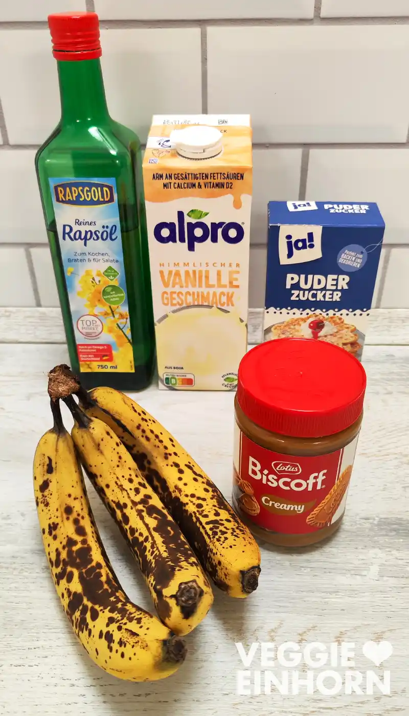 Reife Bananen, Rapsöl, Vanille Sojamilch, Puderzucker und Lotus Biscoff Creme stehen als Zutaten für ein veganes Spekulatius Bananenbrot bereit.