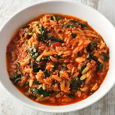 Veganer Orzo-Eintopf mit veganem Hackfleisch, Spinat und Tomaten in einem weißen Teller auf hellem Holztisch.