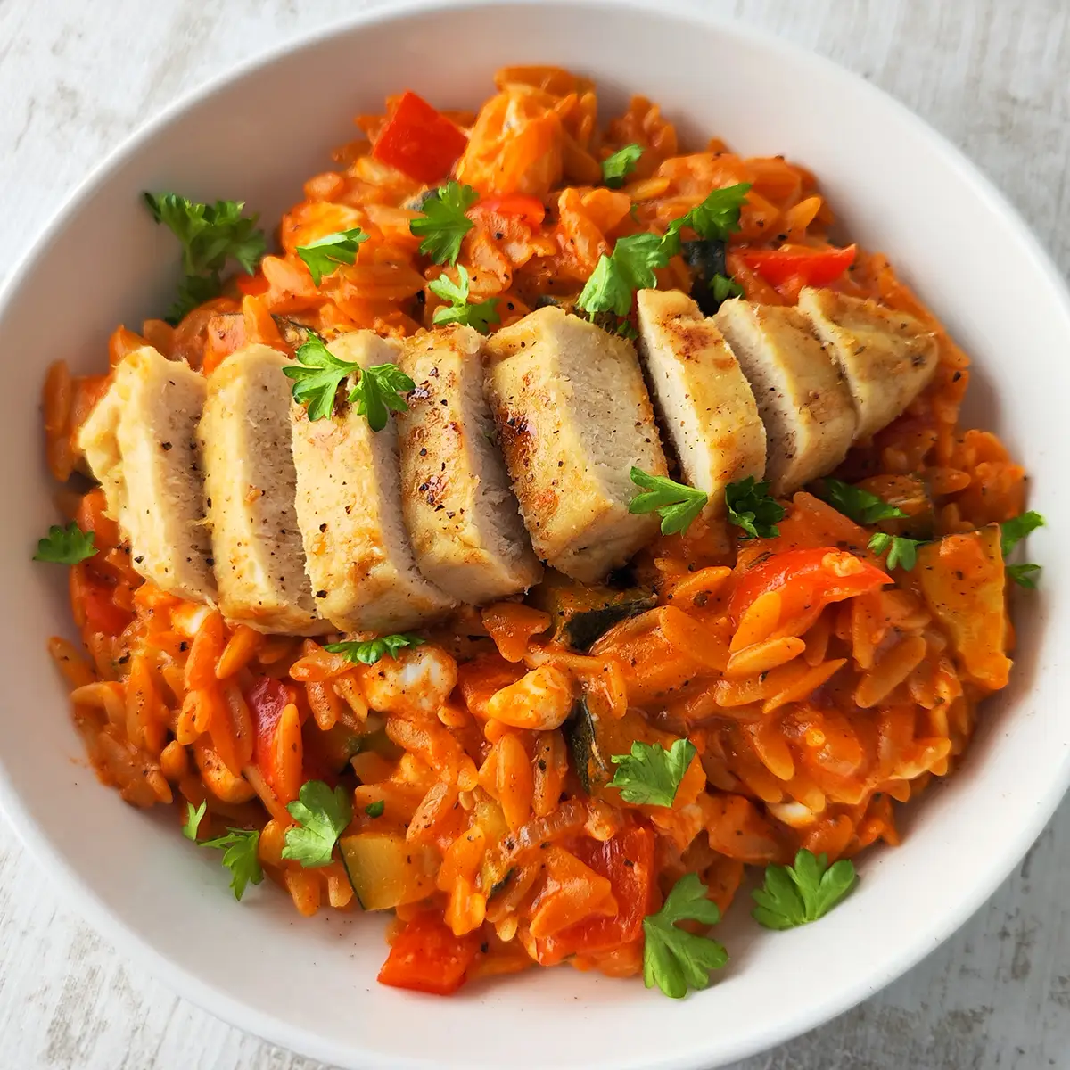 Ein Teller mit Orzo-Pfanne mit Gemüse, dazu in Scheiben geschnittenes veganes Zitronenhähnchen und frische Petersilie als Garnitur.