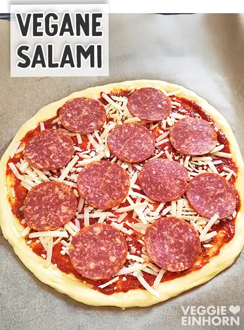 Runder Pizzaboden auf einem Backblech, mit Tomatensauce, veganem Käse und belegt mit Scheiben veganer Salami, darüber Textaufschrift Vegane Salami.