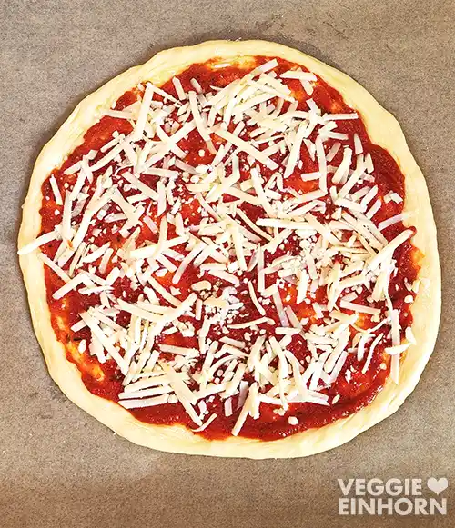 Runder Pizzaboden auf einem Backblech, bestrichen mit roter Pizzasauce und bestreut mit veganem Pizzakäse.