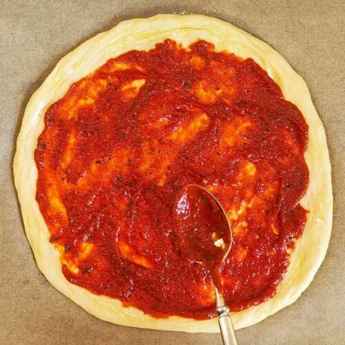 Auf einem runden Pizzateig, der auf Backpapier liegt, wird mit einem Löffel eine vegane Pizzasoße gleichmäßig verteilt.