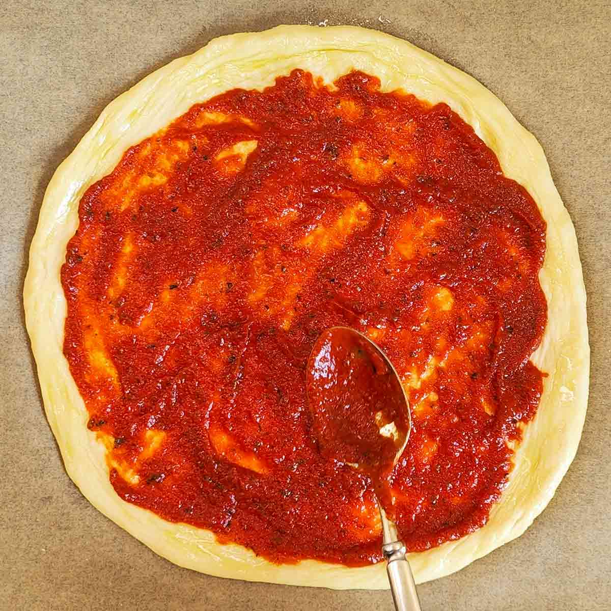 Auf einem runden Pizzateig, der auf Backpapier liegt, wird mit einem Löffel eine vegane Pizzasoße gleichmäßig verteilt.