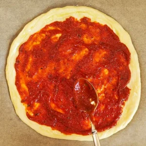 Vegane Pizzasoße wird mit einem Löffel auf einem runden Pizzaboden verteilt.