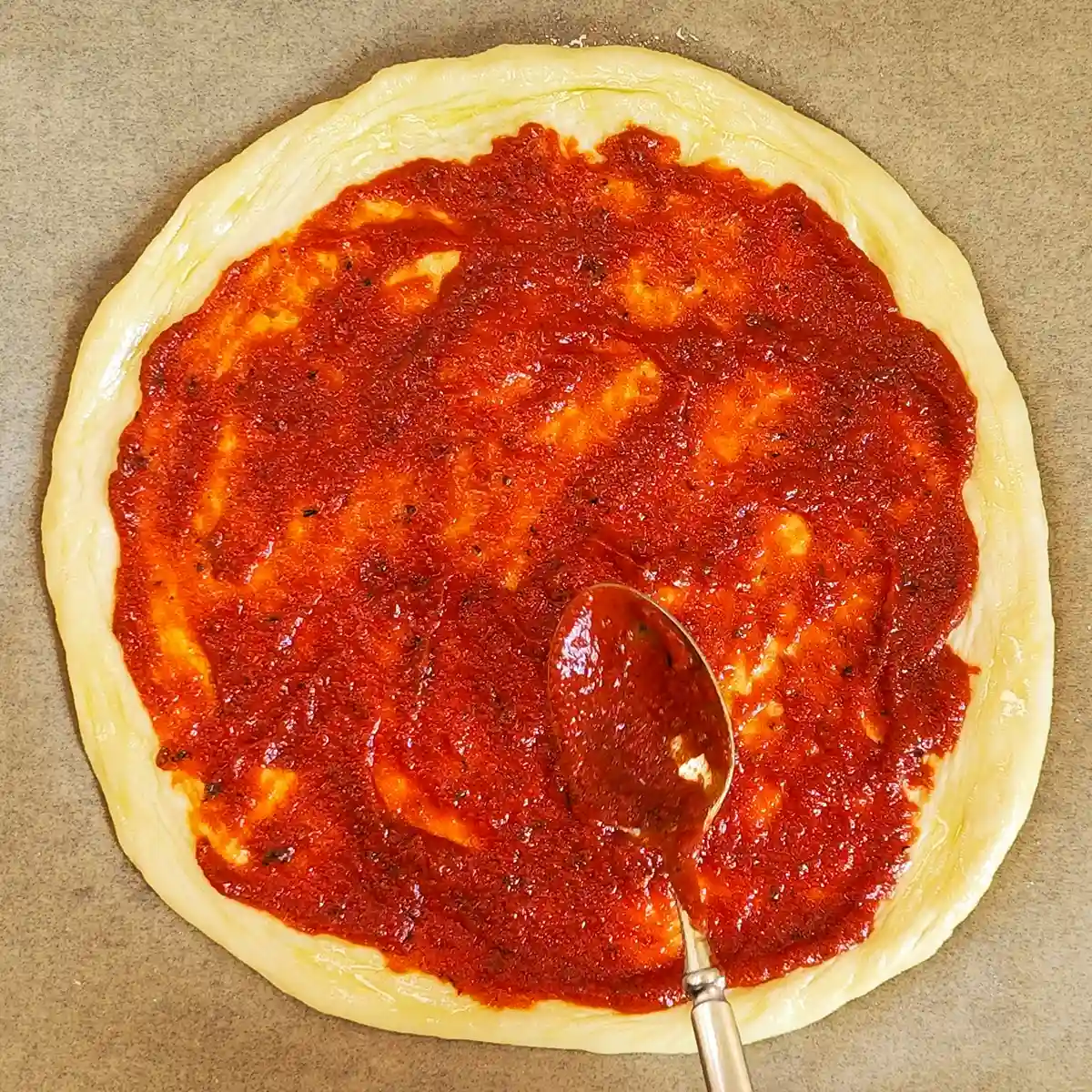Vegane Pizzasoße wird mit einem Löffel auf einem runden Pizzaboden verteilt.