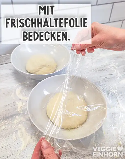 Zwei Schüsseln mit Teigkugeln stehen auf einer bemehlten Arbeitsfläche, zwei Hände decken eine der Schüsseln mit Frischhaltefolie ab. Auf dem Bild steht der Text Mit Frischhaltefolie bedecken.