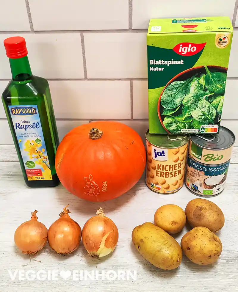 Mehrere Zutaten liegen auf einer hellen Arbeitsfläche, darunter eine Flasche Rapsöl, ein Hokkaidokürbis, Zwiebeln, Kartoffeln, eine Dose Kichererbsen, eine Dose Kokosmilch und eine Packung gefrorener Blattspinat.
