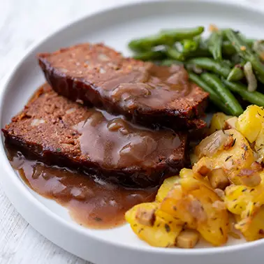 Nahansicht von veganem Braten auf einem weißen Teller mit Bratkartoffeln, Bohnen und Bratensoße als Kategoriebild für vegane Weihnachtsessen.