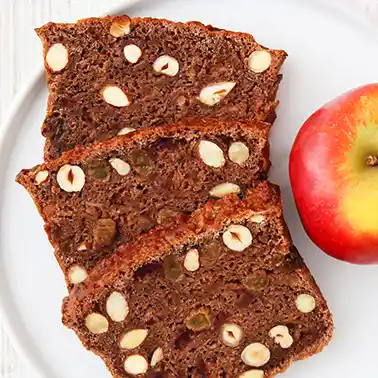 Drei Scheiben veganes Apfelbrot mit Nüssen auf einem weißen Teller mit einem Apfel als Kategoriebild für die Kategorie Rezepte für veganes Weihnachtsgebäck.