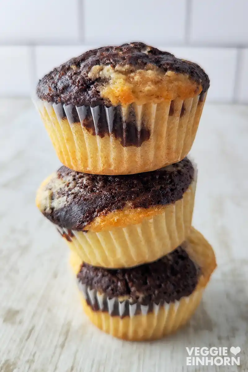 Drei vegane Marmormuffins mit goldgelbem Teig und dunklen Schokoladenwirbeln sind übereinandergestapelt, auf einer hellen Holzoberfläche vor einem hellen Kachelhintergrund.