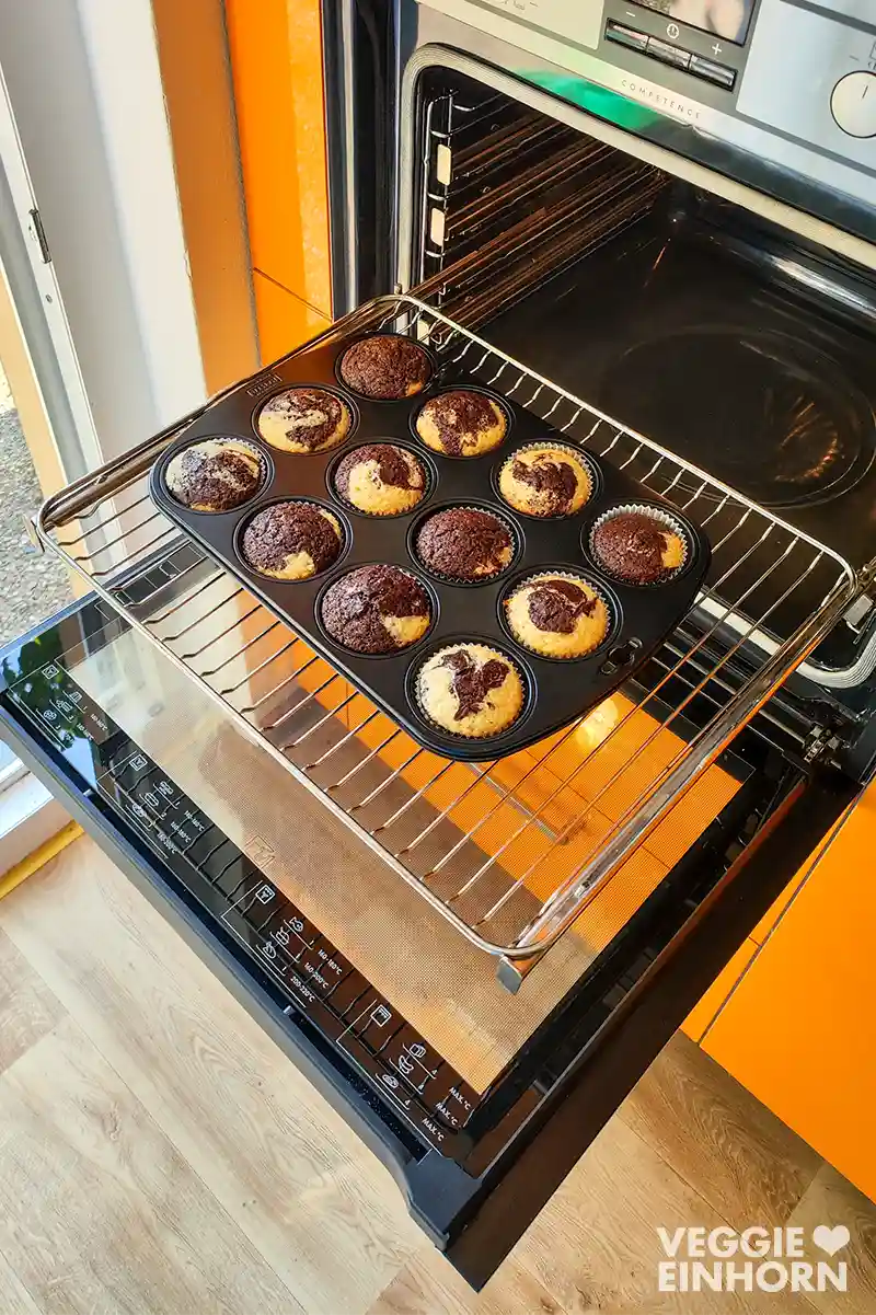 Ein Backofen mit geöffneter Tür, auf dessen Rost ein Muffinblech mit zwölf frisch gebackenen veganen Marmormuffins steht, die goldbraun und marmoriert sind.
