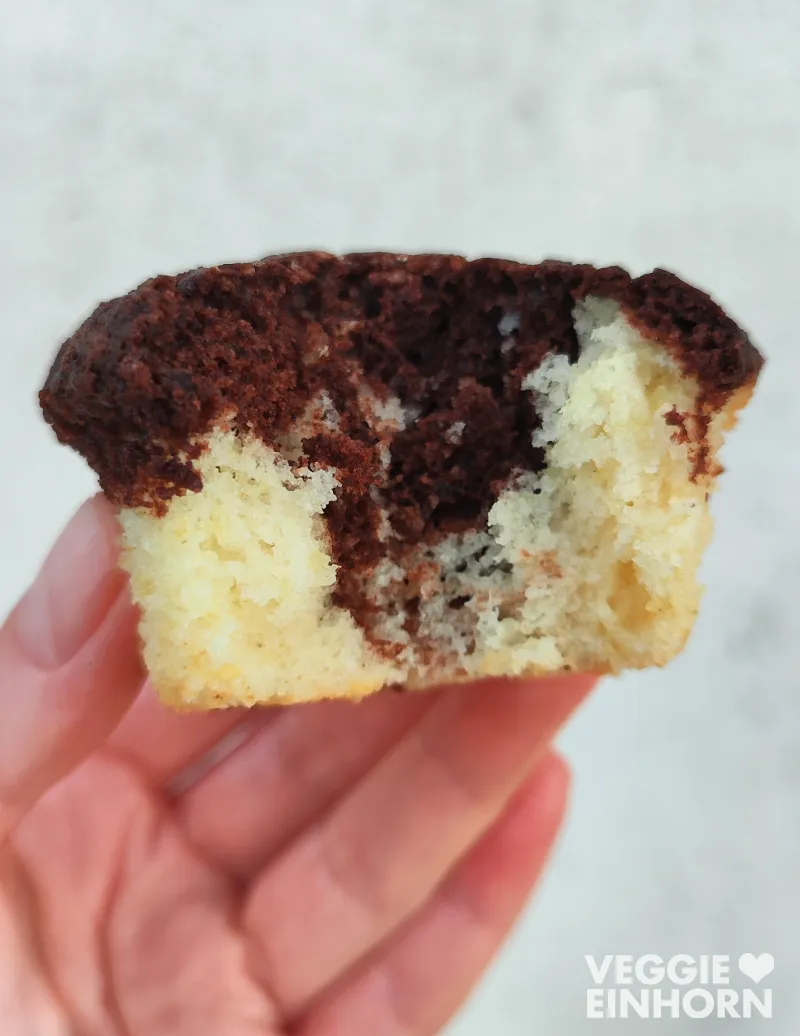 Eine Hand hält einen halbierten veganen Marmormuffin, der innen eine fluffige, marmorierte Struktur aus hellem Vanilleteig und dunklem Schokoladenteig zeigt.