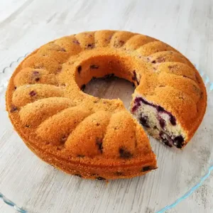 Saftiger veganer Blaubeerkuchen in Rohrbodenform mit herausgeschnittenem Stück auf einer Kuchenplatte aus Glas.