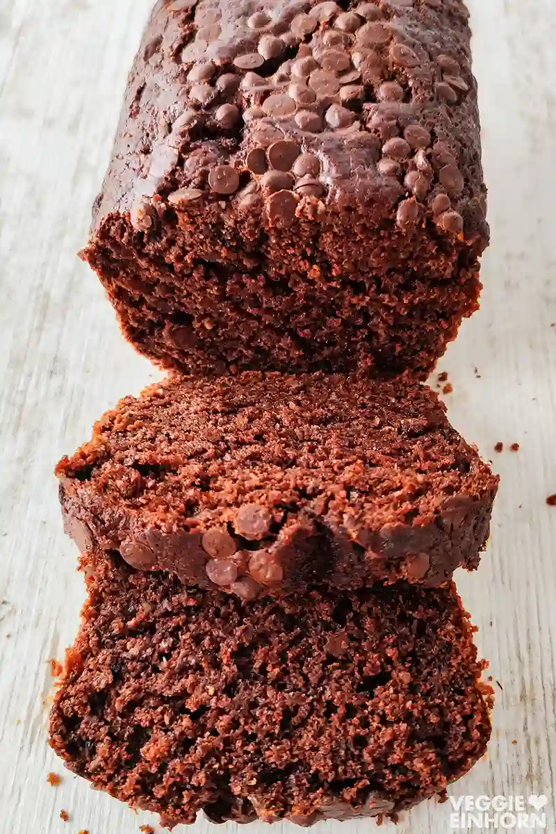 Angeschnittener veganer Schokokuchen mit Schokotröpfchen, zwei Kuchenstücke liegen davor auf einem hellen Holztisch.