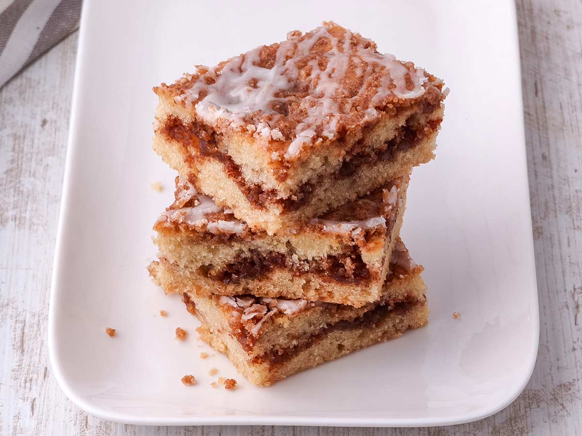 Mehrere übereinandergestapelte Stücke von Zimtkuchen mit Zuckerguss auf einer Kuchenplatte.